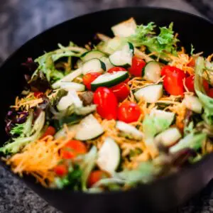 A black bowl filled with a mixed salad containing lettuce, shredded cheese, sliced cucumbers, grape tomatoes, and onions.