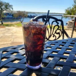 A glass of iced dark soda with a straw sits on a black metal outdoor table, overlooking a lake and trees on a sunny day.