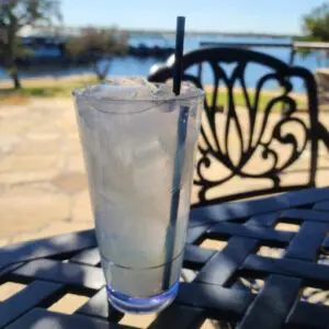 A clear plastic cup filled with ice and a light-colored drink with a straw, placed on a metal patio table outdoors near a body of water.