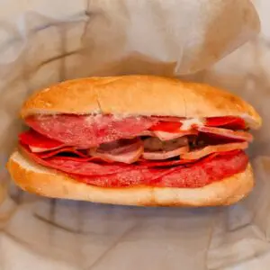 A sandwich on a crusty roll filled with layers of salami, pepperoni, other deli meats, pickles, and mayonnaise, wrapped in parchment paper.