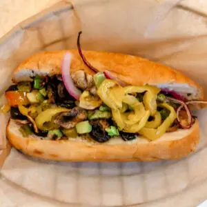 A sandwich roll filled with sliced onions, black olives, banana peppers, tomatoes, cucumbers, and green peppers, served on parchment paper.