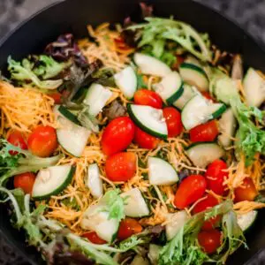 A bowl of salad with shredded cheese, cherry tomatoes, sliced cucumbers, and mixed leafy greens.