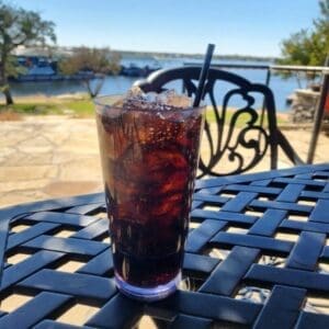 A glass of iced dark soda with a straw sits on a black metal outdoor table, overlooking a lake and trees on a sunny day.
