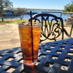 A glass of iced tea with a straw sits on a black metal patio table, with a lake and trees visible in the background.