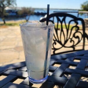 A clear plastic cup filled with ice and a light-colored drink with a straw, placed on a metal patio table outdoors near a body of water.