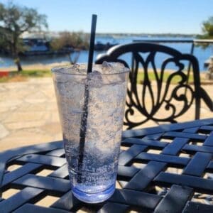 A clear glass filled with ice and sparkling water with a black straw sits on a metal outdoor table, with a lake and trees in the background.