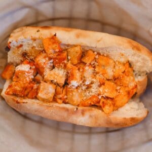 A sandwich roll filled with seasoned, cubed tofu and sprinkled with grated cheese, placed on a paper-lined surface.