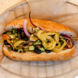 A sandwich roll filled with sliced onions, black olives, banana peppers, tomatoes, cucumbers, and green peppers, served on parchment paper.