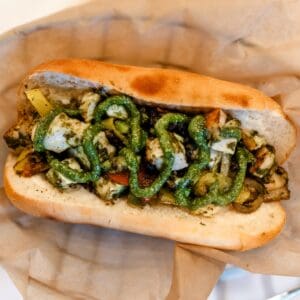 A sandwich roll filled with grilled vegetables, pieces of mozzarella cheese, and drizzled with green pesto sauce, placed on brown parchment paper.