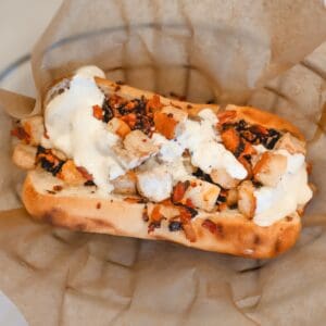 A toasted sandwich roll filled with pieces of grilled chicken and topped with a creamy white sauce, served on brown parchment paper.